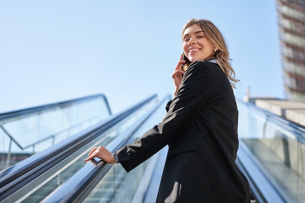 Foto gratuita retrato de mujer de negocios en traje negro subiendo escaleras mecánicas hablando por teléfono móvil vendedora wal