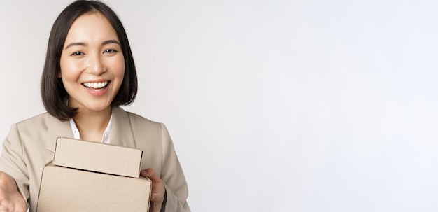 Retrato de mujer de negocios vendedora asiática señalándote dando cajas con órdenes de pie en traje sobre fondo blanco.