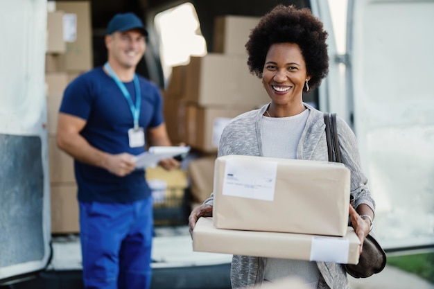 Retrato de una mujer negra feliz que se siente satisfecha con el anuncio de entrega de paquetes sosteniendo cajas mientras mira a la cámara El mensajero está en segundo plano