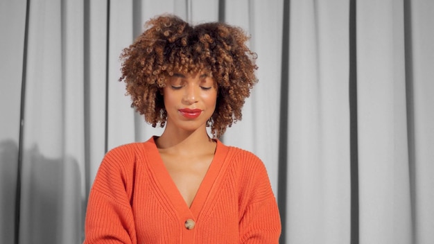 Retrato de una mujer negra de raza mixta con cabello afro rizado texturizado en una chaqueta naranja brillante con maquillaje natural para tonos de piel oscuros mirando hacia abajo