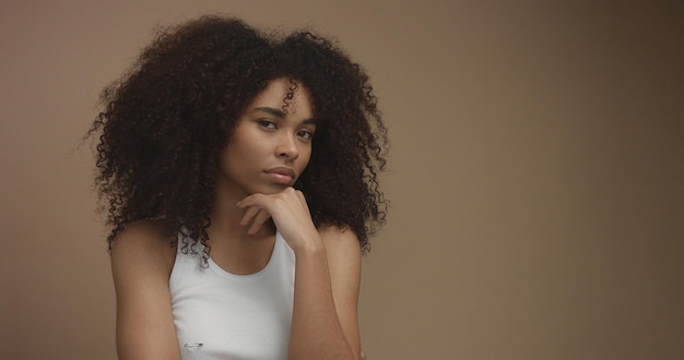 Retrato de mujer negra de raza mixta con gran cabello afro rizado en fondo beige pensando en algo con una mano en la barbilla