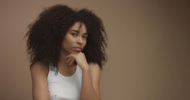 Retrato de mujer negra de raza mixta con gran cabello afro rizado en fondo beige Pensando en algo con una mano en la barbilla