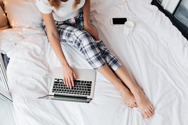 Retrato de mujer en pantalones a cuadros usando laptop. Foto interior de mujer freelance en pijama trabajando en la cama.