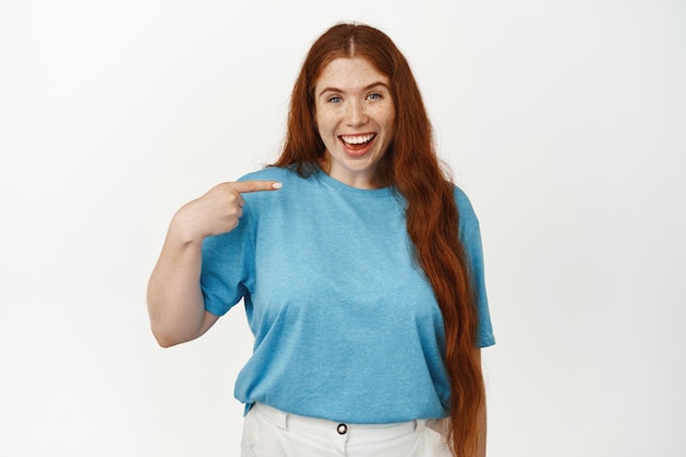 Retrato de mujer pelirroja emocionada jadeando sorprendida y asombrada, señalándose a sí misma y sonriendo divertida, siendo elegida, ganando premio, de pie sobre fondo blanco.