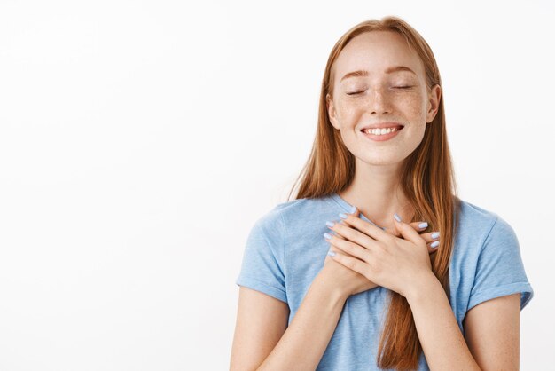 Retrato de mujer pelirroja emocional feliz soñadora con pecas cerrando los ojos y sonriendo alegre y relajado tomados de la mano en el pecho