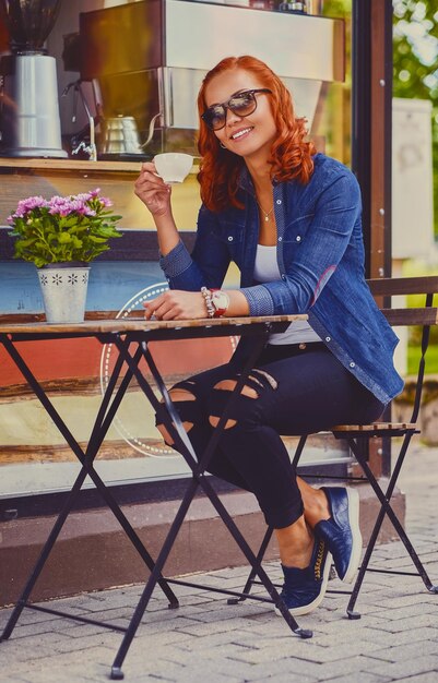 Retrato de mujer pelirroja con gafas de sol, bebe café en una cafetería en una calle.