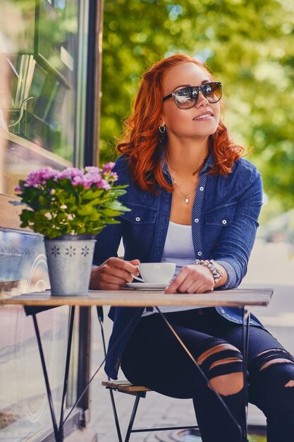 Retrato de mujer pelirroja con gafas de sol, bebe café en una cafetería en una calle.