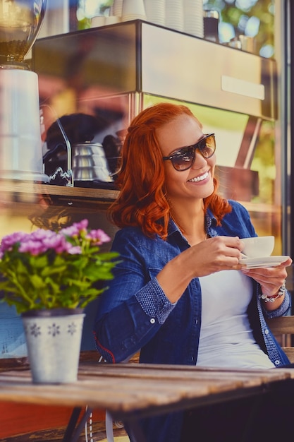 Retrato de mujer pelirroja con gafas de sol, bebe café en una cafetería en una calle.