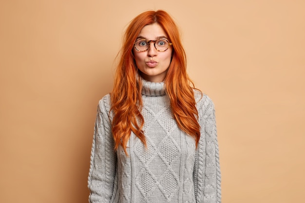 Retrato de mujer pelirroja guapa mantiene los labios redondeados tiene expresión romántica en la cara quiere besar a alguien viste suéter gris