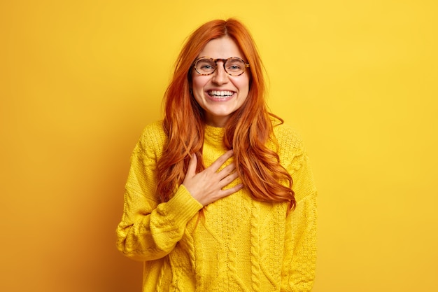 Retrato de mujer pelirroja llena de alegría sonríe ampliamente mantiene la mano en el pecho sonríe ampliamente expresa buenas emociones se siente complacido usa lentes transparentes suéter casual.