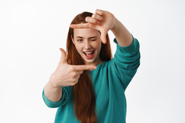 Retrato de mujer pelirroja mira a través de marcos de manos con ojo creativo, mide algo, busca el lugar perfecto para la toma, de pie sobre fondo blanco