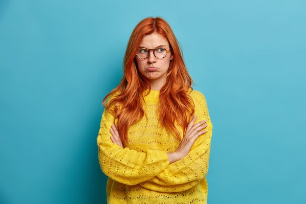 Retrato de mujer pelirroja ofendida triste cruza los brazos y frunce los labios tristes después de romper o pelear con el novio reacciona a las palabras negativas tiene expresión de cara molesta vestida con suéter amarillo.