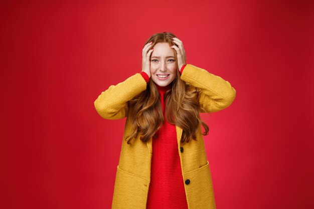 Retrato de mujer pelirroja en pánico cogidos de la mano apretados a la cabeza apretando los dientes y frunciendo el ceño desconcertado y preocupado, de pie ansioso y preocupado volviéndose loco sobre fondo rojo.