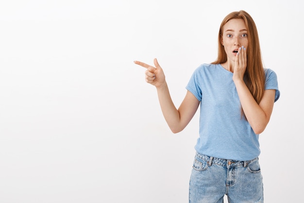 Retrato de mujer pelirroja preocupada y aturdida que expresa empatía y preocupación apuntando hacia la izquierda con el brazo estirado que cubre abierto de la boca sacudida