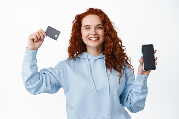 Retrato de mujer pelirroja satisfecha con cabello rizado, sonriendo complacida, mostrando la pantalla vacía del teléfono inteligente y la tarjeta de crédito plástica, de pie en blanco