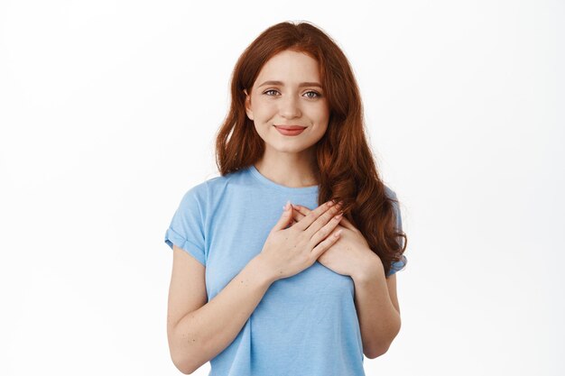 Retrato de mujer pelirroja sonriente esperanzada, tomándose de la mano en el corazón y mirando tiernamente a la cámara, siendo tocada, decir gracias, complacida y sincera, de pie contra el fondo blanco