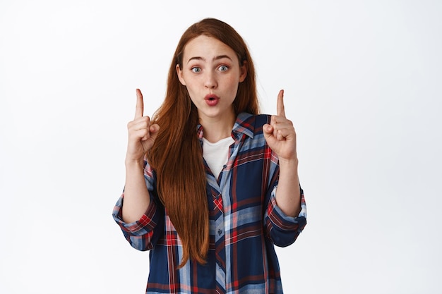 Retrato de mujer pelirroja sorprendida, señalando un increíble trato promocional, mostrando publicidad, anuncio fantástico, de pie contra un fondo blanco.