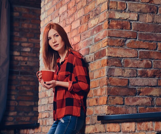 Retrato de mujer pelirroja vestida con una camisa de lana roja y jeans sobre la pared de ladrillos rojos.