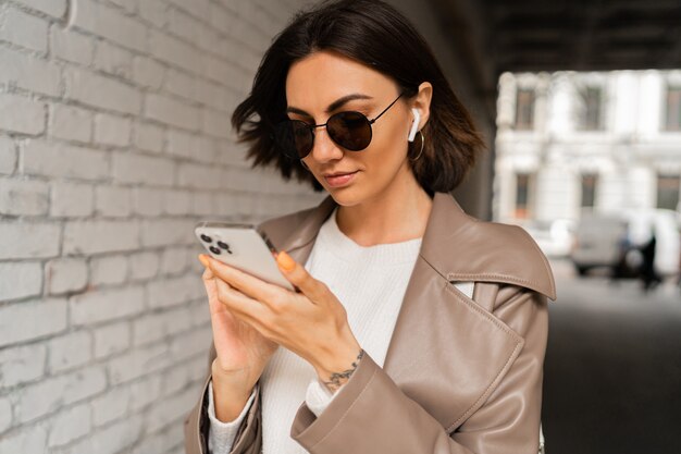 Retrato de mujer de pelo corto elegante con auriculares en abrigo de cuero casual y gafas de sol de cerca con smartphone y posando sobre pared de ladrillo urbano de cerca