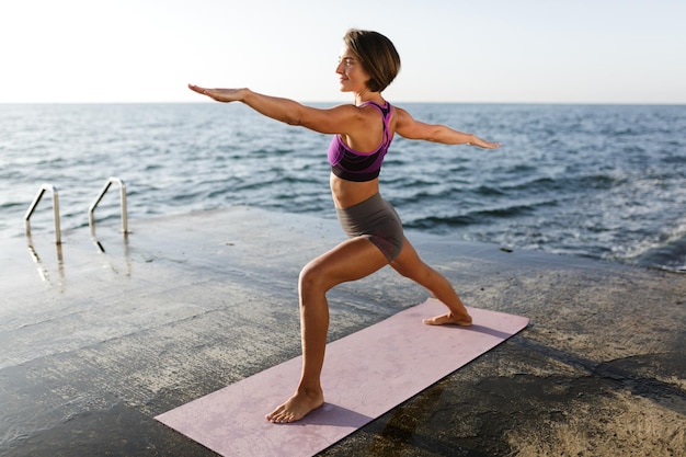 Retrato de mujer con pelo corto oscuro de pie y tirando de las manos a un lado mientras entrena poses de yoga junto al mar. Señorita en top deportivo y pantalones cortos practicando yoga con mar de fondo
