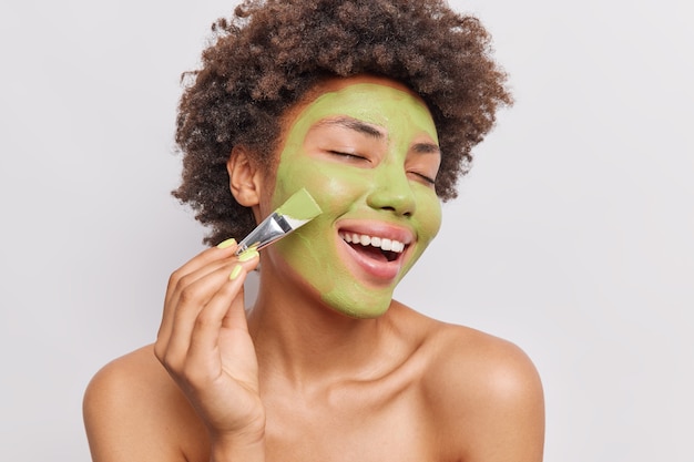 Retrato de mujer de pelo rizado aplica mascarilla nutritiva verde con cepillo cosmético