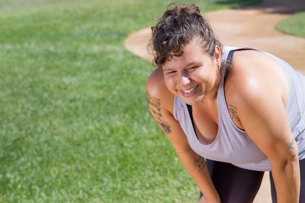 Retrato de mujer con pelo rizado haciendo deporte en un día soleado. Mujer gordita en top gris con tatuajes mirando hacia los lados. Deporte, concepto de cuerpo positivo