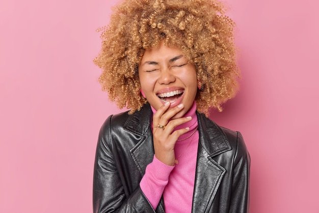 El retrato de una mujer de pelo rizado muy contenta se ríe alegremente mantiene los ojos cerrados escucha algo muy divertido lleva una chaqueta de cuero aislada sobre un fondo rosa. Las emociones de las personas y el concepto de estilo.