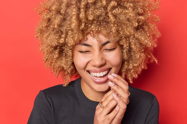 El retrato de una mujer de pelo rizado se ríe alegremente, mantiene los ojos cerrados, sonríe con dientes, mantiene las manos juntas vestidas con una camiseta negra informal aislada sobre un fondo rojo vivo, escucha algo divertido