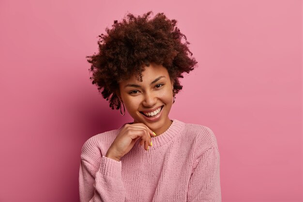 Retrato de mujer de pelo rizado toca la barbilla, inclina la cabeza, sonríe y mira, vestida con un suéter, expresa positividad y alegría, viste un suéter informal, aislado sobre una pared rosa pastel