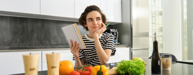 Retrato de una mujer pensante con un cuaderno de cocina escribiendo los ingredientes de la receta decidiendo una comida