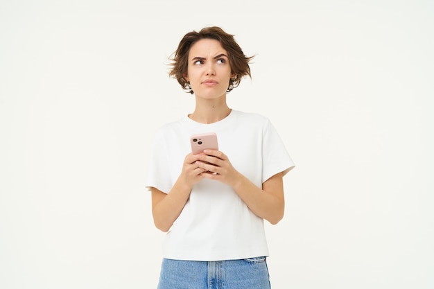 Foto gratuita retrato de una mujer pensante con un teléfono inteligente que parece perplejo y reflexiona sobre la toma de decisiones de pie