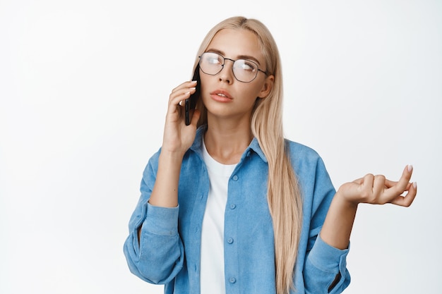 Retrato de mujer pensativa con gafas hablando por teléfono móvil y encogiéndose de hombros, mirando perplejo durante una llamada, de pie sobre blanco