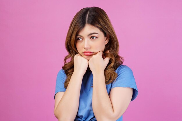 Retrato de una mujer pensativa, miserable y abatida, con una camiseta informal, manteniendo las manos bajo la barbilla, aislada sobre un fondo rosado