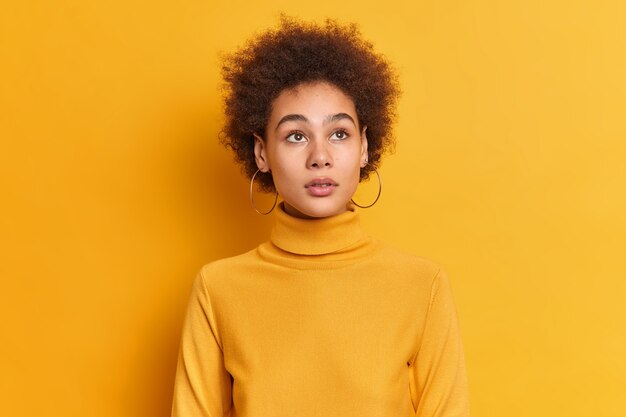 El retrato de una mujer pensativa de pelo rizado concentrada en algún lugar tiene una belleza natural y lleva un cuello alto informal y pendientes.