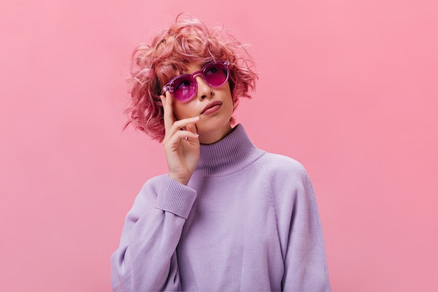 Retrato de mujer pensativa rizada de pelo rosa en suéter púrpura y gafas de sol fucsia posando en la pared aislada