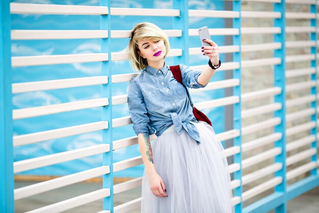 Retrato de una mujer de pie con el pelo corto y rubio, labios rosados brillantes y maquillaje desnudo haciendo selfie