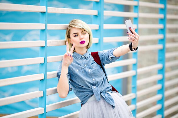 Retrato de una mujer de pie con el pelo corto y rubio, labios rosados brillantes y maquillaje desnudo haciendo selfie