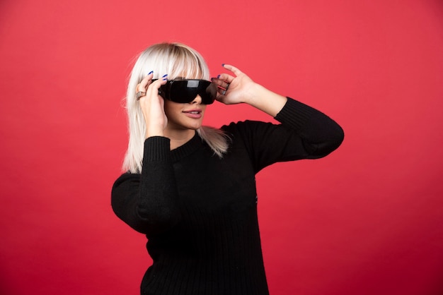 Retrato de mujer de pie y posando con gafas negras sobre una pared roja.