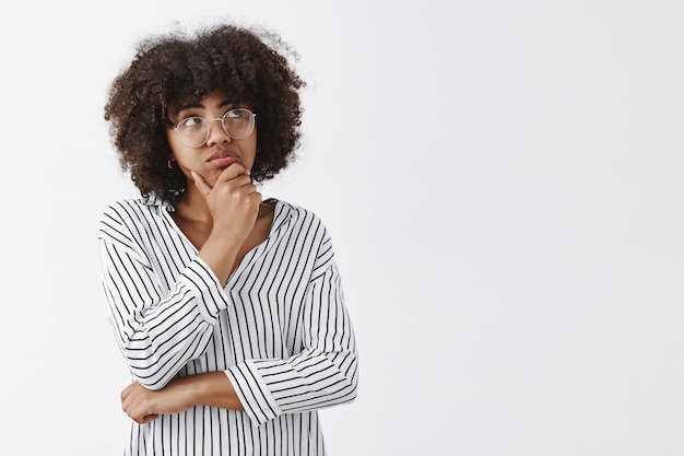 Retrato de mujer de piel oscura linda soñadora y enfocada pensativa con el pelo rizado haciendo pucheros sosteniendo la mano en la barbilla y mirando la esquina superior derecha mientras piensa y hace una idea o plan