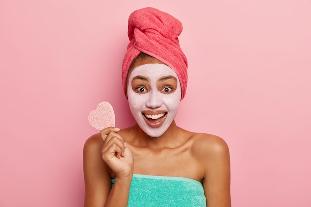 Retrato de mujer de piel oscura sonriente feliz aplica mascarilla de barro, tiene tratamiento de rejuvenecimiento, sostiene una esponja para quitar el maquillaje, tiene expresión de alegría