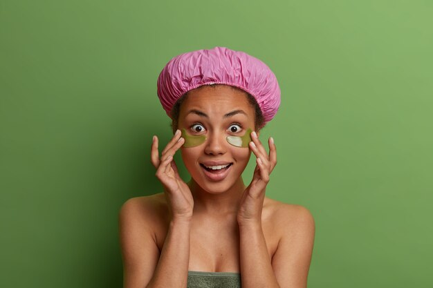 Retrato de mujer de piel oscura sorprendida que toca la piel suave, se aplica parches en los ojos para reducir la hinchazón y las líneas finas, usa gorro de baño, una toalla suave alrededor del cuerpo, aislado sobre una pared verde.