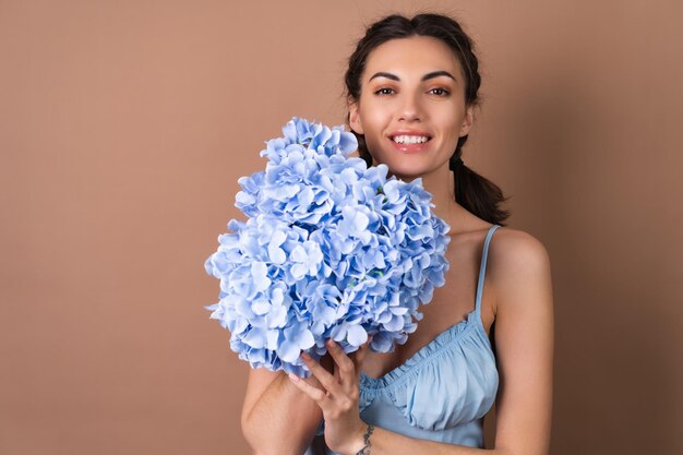 Retrato de una mujer con piel perfecta y maquillaje natural sobre un fondo beige con coletas en un vestido con un ramo de flores azules