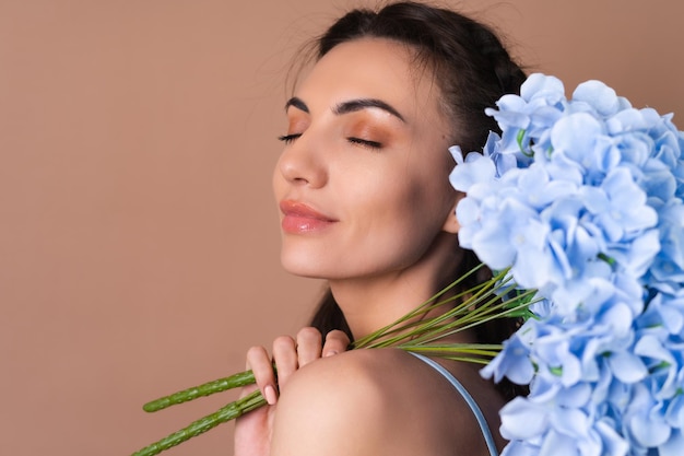 Retrato de una mujer con piel perfecta y maquillaje natural sobre un fondo beige con coletas en un vestido con un ramo de flores azules
