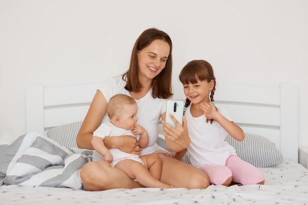 Retrato de mujer positiva con camiseta blanca de estilo casual sentada en la cama con dos hijas, sosteniendo el teléfono inteligente en las manos, haciendo videollamadas o transmitiendo en vivo.