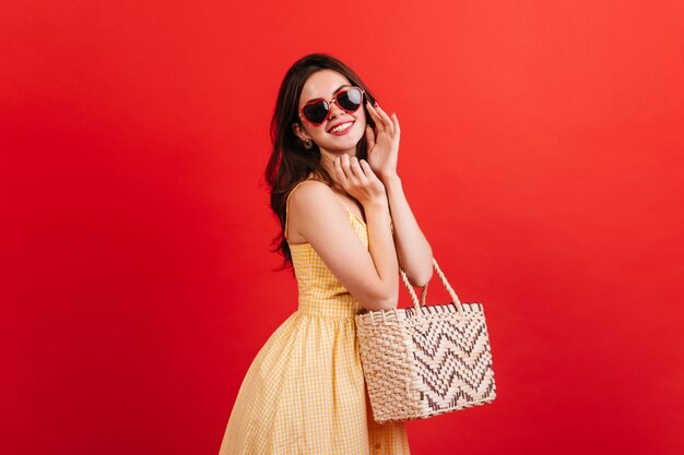 Retrato de mujer positiva de muy buen humor posando en la pared roja. Señora de pelo oscuro en traje de verano brillante con bolsa de playa.