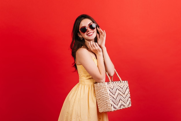 Retrato de mujer positiva de muy buen humor posando en la pared roja. Señora de pelo oscuro en traje de verano brillante con bolsa de playa.