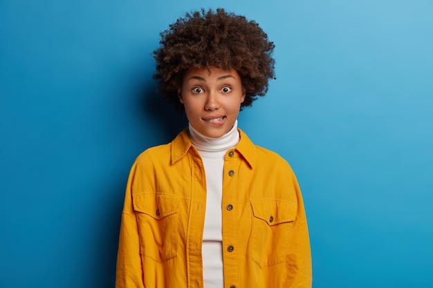 Retrato de mujer preocupada de piel oscura muerde los labios y mira sorprendentemente a la cámara, vestida con cuello alto blanco y camisa amarilla, nerviosa, aislada en azul