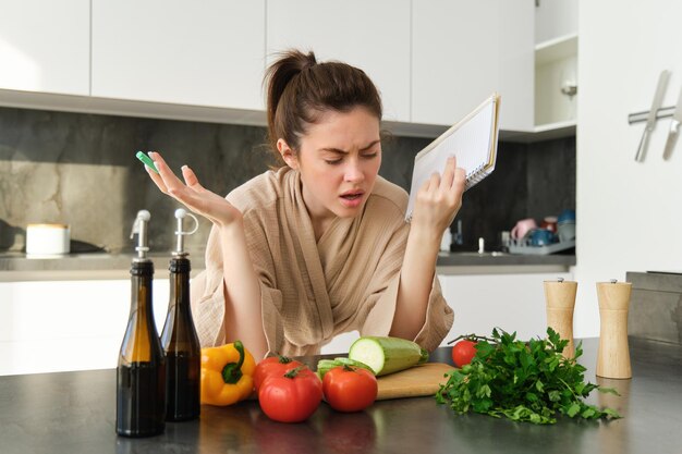 Retrato de una mujer que no sabe cocinar y parece confundida mientras prepara la comida sosteniendo un libro de recetas revisando la tienda de comestibles