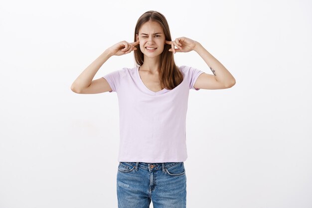 Retrato de mujer que siente malestar por el ruido fuerte que cubre los oídos con los dedos índices entrecerrando los ojos y apretando los dientes cerrando los ojos mientras escucha un fuerte golpe