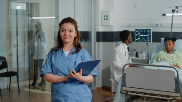 Retrato de mujer que trabaja como enfermera con papeles de chequeo mientras está de pie en la sala del hospital. Asistente médico con documentos mirando a cámara y sonriendo, dando ayuda al especialista.